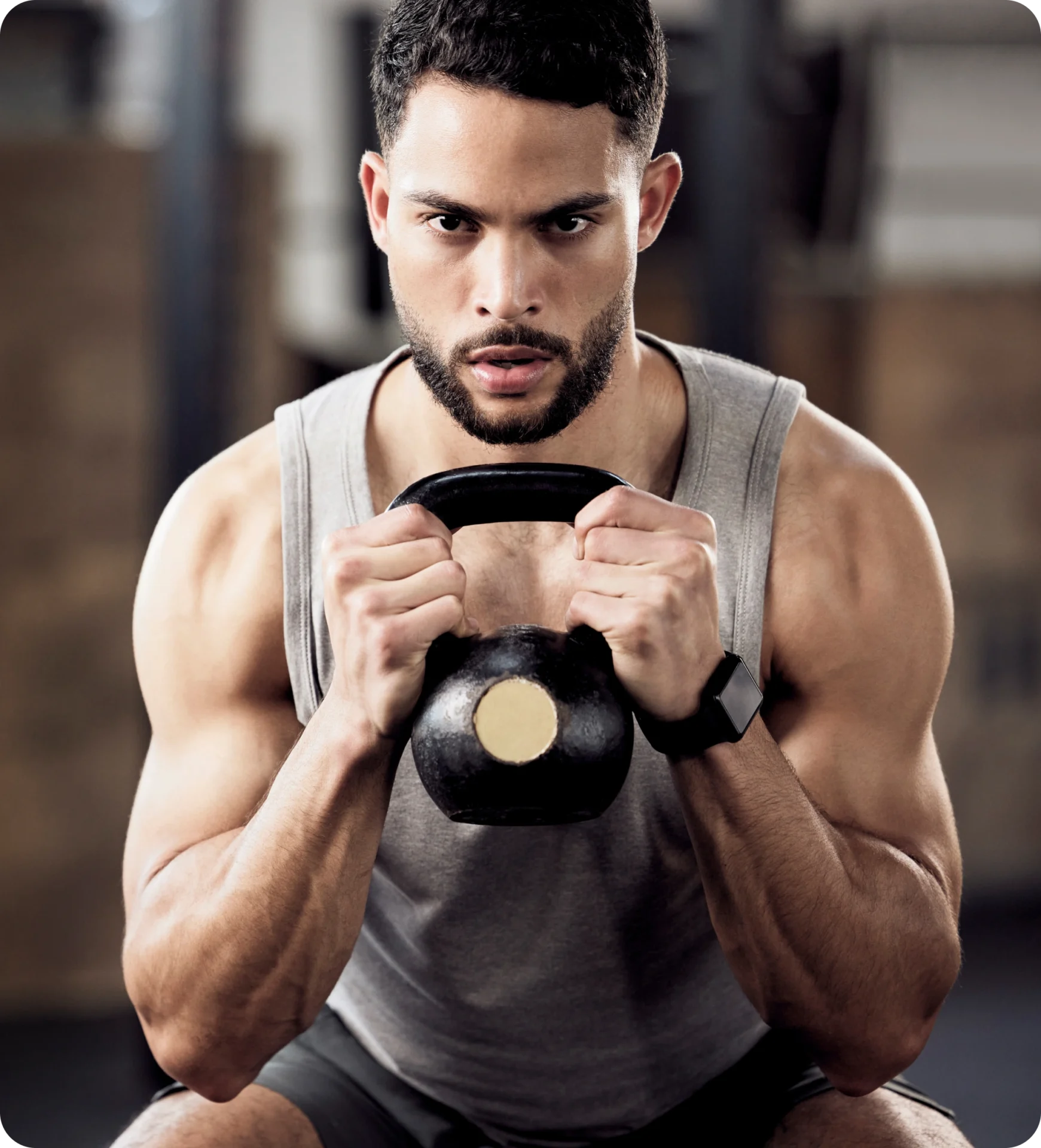 A person holding a kettlebell with both hands during a full squat workout, wearing a gray tank top and a wristwatch.